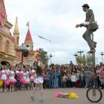 A jugger balancing on a tall "giraffe" unicycle passing juggling clubs with girl from a big audience during show at amusement park